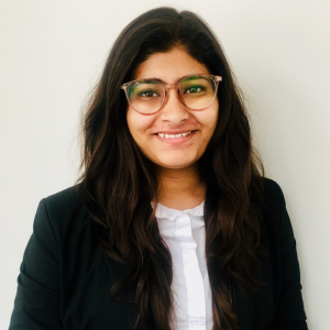 Portrait of Riya in a white shirt and black blazer, smiling against a white background.