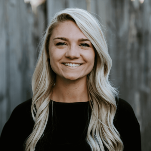 Blonde woman with a black shirt and a smile standing in front of a fence.