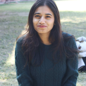 An outdoor portrait of a woman in a green sweater with a building and landscaping in the background.