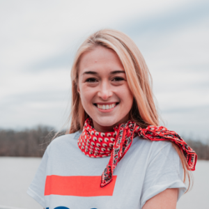 A smiling blonde woman wearing a gray t-shirt with orange stripe and a red scarf.