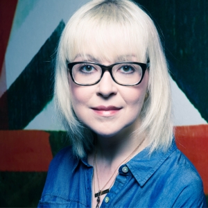 A blonde woman with black glasses and a blue shirt smiling for a photo in front of a painting.