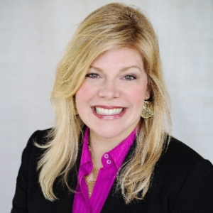 A woman with blonde hair wearing a pink shirt and black blazer smiling for a picture.