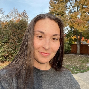 Devyn Rheuby stands in front of a tree in a park wearing a gray t-shirt.
