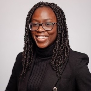 A woman with glasses and braids wearing a black turtleneck and blazer poses for a photo.