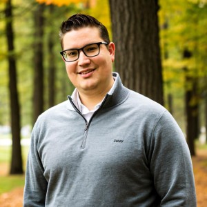 A man with glasses and a gray sweatshirt stands smiling in a forest with yellow leaves.