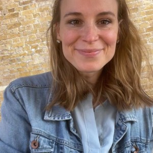 Smiling woman with brown hair wearing a denim jacket standing in front of a brick wall.