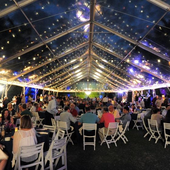 Groups of people sit around tables at an outdoor event under a tent