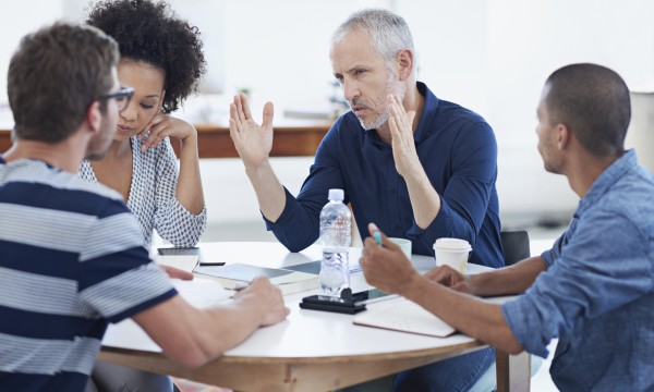 Three people sitting around a table with a man raising his hand in the middle of the table.