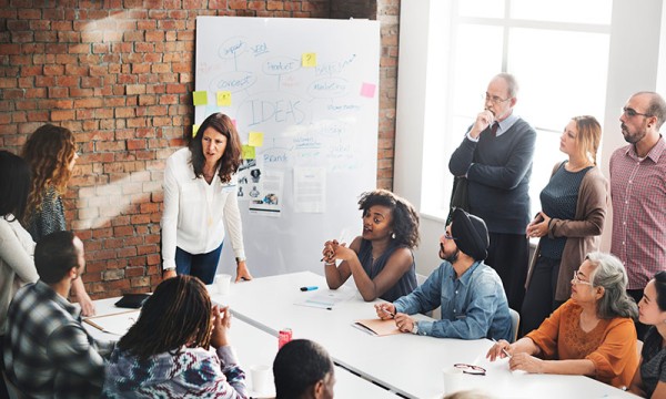 Group of people sitting around a table while a woman is standing and talking to them.