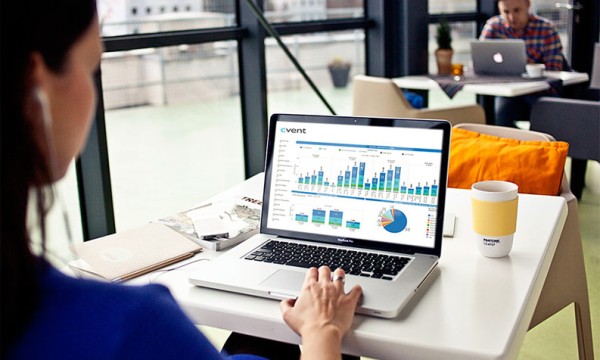Woman using a laptop at a desk with a graph on the screen and man in background.