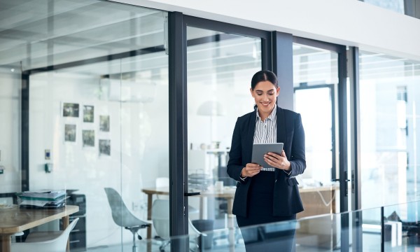 A woman in a suit holding a tablet in an office with glass walls and doors.