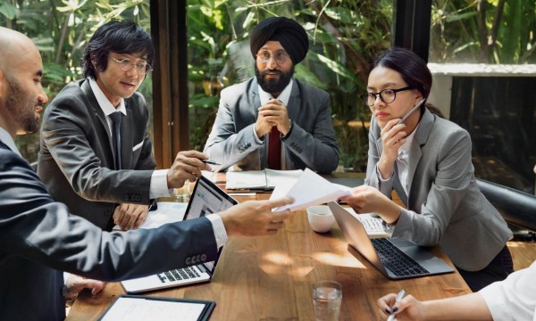 Businesspeople in a meeting with laptops, documents, and a glass window overlooking greenery.