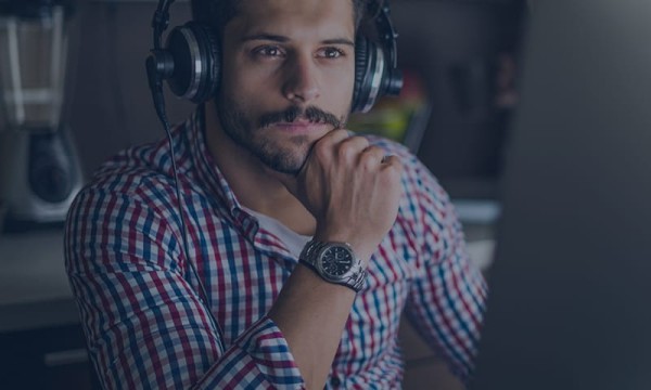 A man in a red checkered shirt wearing headphones is thinking while looking at his computer screen.