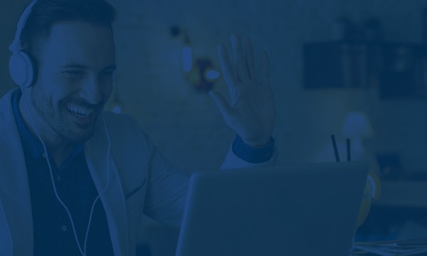 A man with headphones on, sitting at a laptop, smiling, and waving his hand.