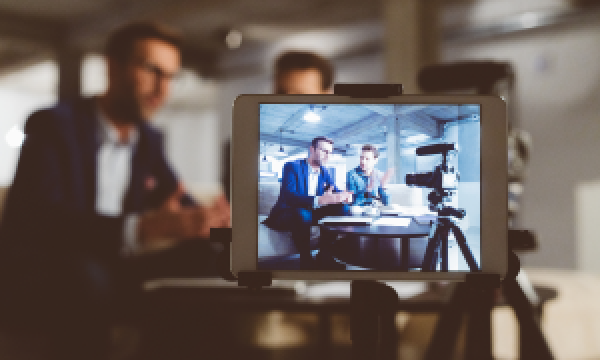 A man is recording a video of two men sitting in front of a desk.