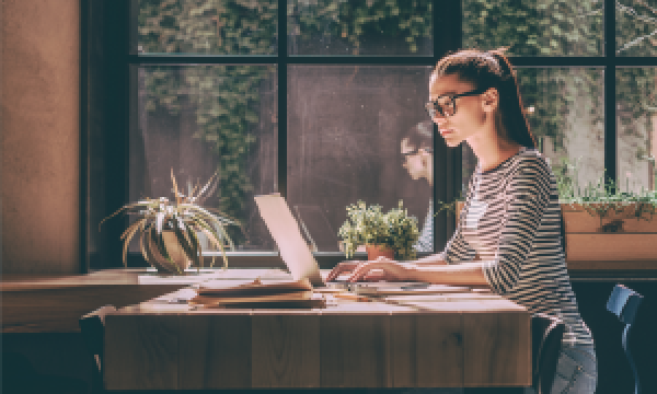 Woman with glasses working on a laptop at a desk by a window with plants outside.