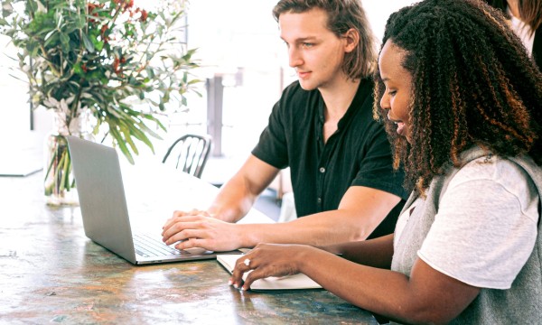 Two people work together on a laptop in a bright, inviting room with flowers and a wooden table.