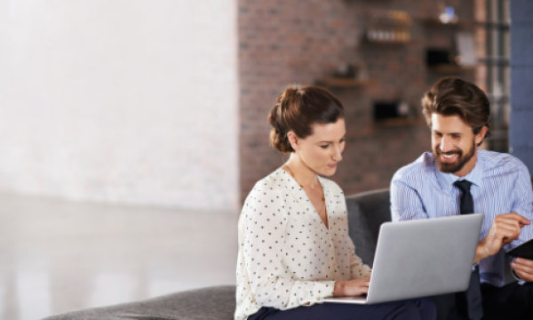 A man and woman sit on a couch, smiling while working on a laptop together.