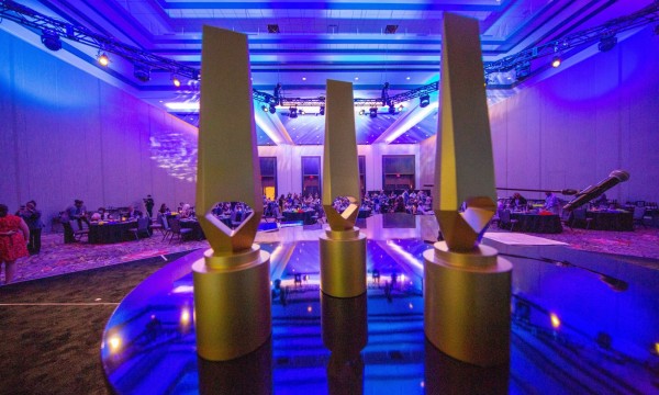 Gold awards stand on a blue table in front of a crowd in a purple lit room.
