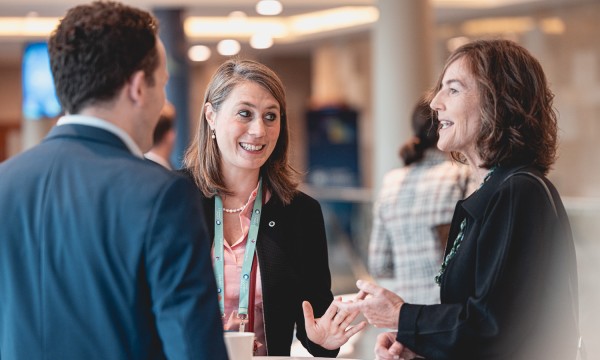 Two women and a man converse at a round table at a conference with others in the background.