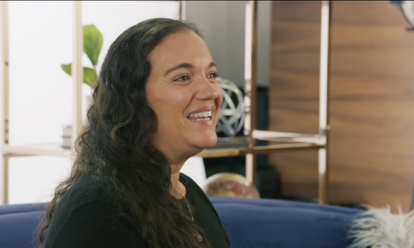 A smiling woman with long hair sits on a blue couch with shelves and plants in the background.