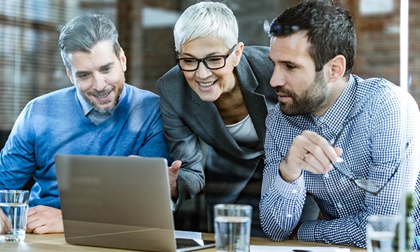 Three business people looking at a laptop on a table with glasses of water in front of them.