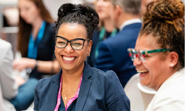Two women laughing at a conference with others in the background.