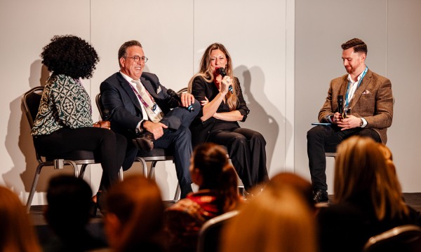 Three people sit on stage, talking into microphones in front of an audience at a conference.