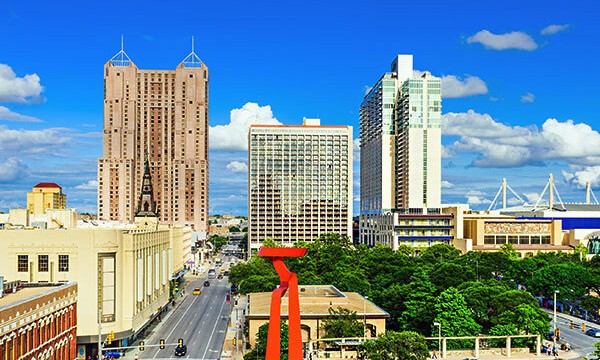 Downtown San Antonio with a blue sky, buildings, a red sculpture, trees, and a road with cars.