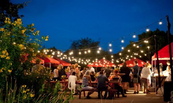 Group of people at tables and benches at night in the garden with glowing string lights.