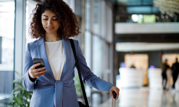 Woman in business attire standing in airport holding her phone and a rolling suitcase.