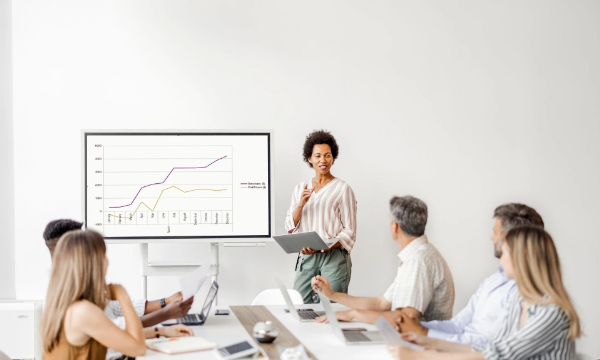 A group of people sitting at a table with a woman standing and talking in front of them.