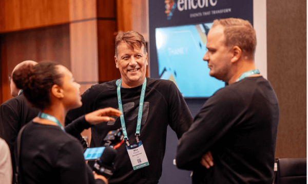Three people at a conference with a large "Encore" sign in the background, and a table and chairs nearby.