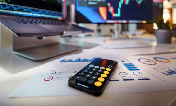 A desk with a remote, documents, a laptop, and a computer with a stock chart on the screen.