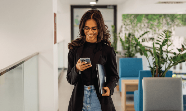 Woman in office corridor with smartphone and binder, surrounded by plants and modern seating.