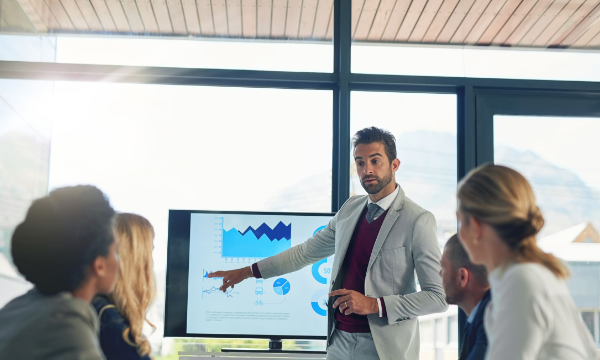 A man presents data on a screen to a group of people in a conference room.