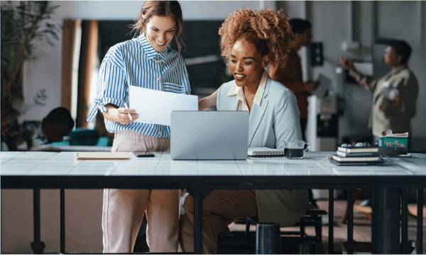 Two women are laughing and reviewing documents with a laptop in front of them in a casual office environment.