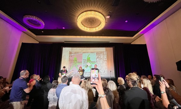 A group of people are standing and raising their hands in a hall with a projector screen.