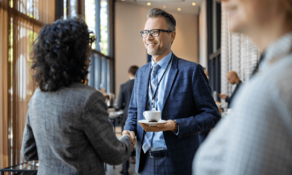 A businessman greeting a woman while holding a cup and wearing a lanyard and glasses.
