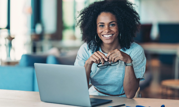 A woman is sitting at a desk with a laptop, smiling and holding a pair of glasses.