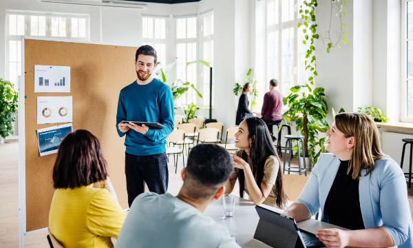 A group of people in a room with a board and plants, having a meeting.