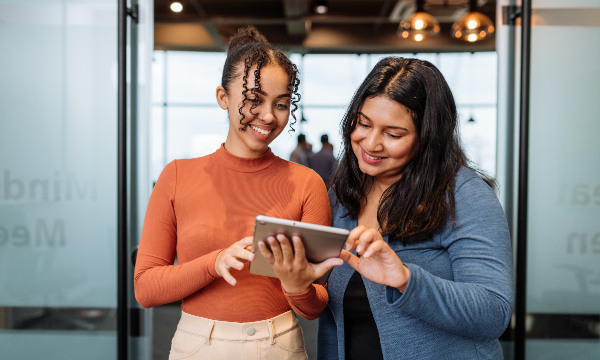 Two women are smiling while looking at a tablet in front of a glass wall at Microsoft.