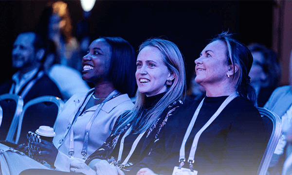 Three women laughing at an event, with one holding a coffee cup and others in the background.