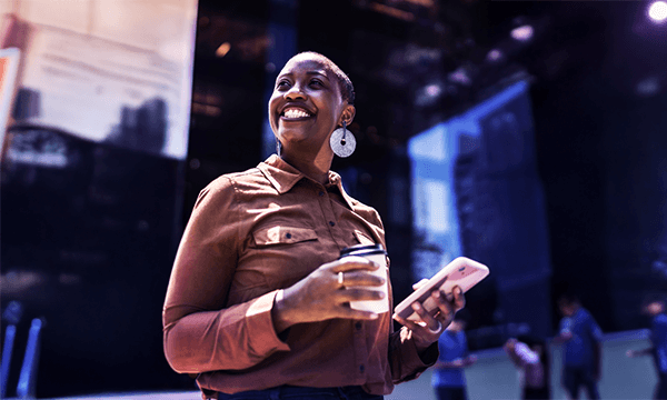 A person in a brown shirt holds a coffee cup and a smartphone, standing in a modern indoor space