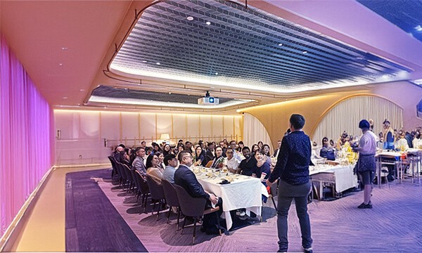 A speaker addresses an audience seated at tables in a modern banquet hall