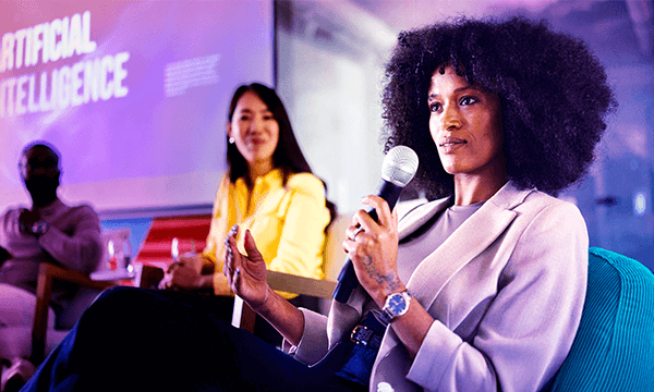 A woman speaks passionately into a microphone on stage, with a colorful backdrop displaying "Artificial Intelligence."