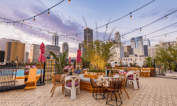 Rooftop dining area with vibrant decor, overlooking a city skyline at sunset, string lights above, and tropical plants around.