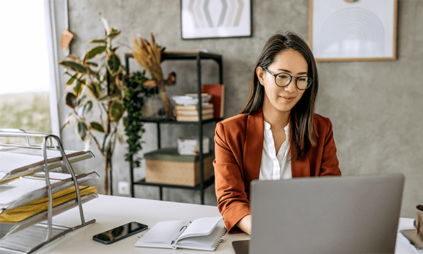 A woman in a brown blazer sits at a desk with a laptop, stationery, and plants, in a modern, well-lit office space.