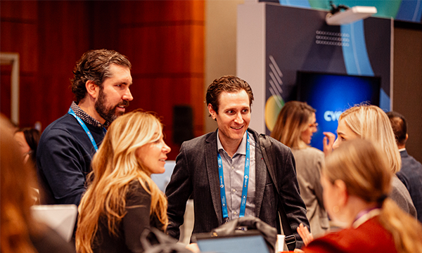 A group of professionals engage in conversation at a conference, with promotional banners and a laptop visible in the background.