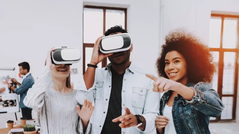 Three people with VR goggles looking at something and pointing, with two people behind them in an office.